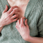 Close-up of a person in a green sweater scratching red, irritated skin on their chest.