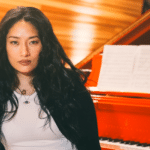 Chloe Flower seated in front of a red grand piano with sheet music, wearing a black jacket over a white top.